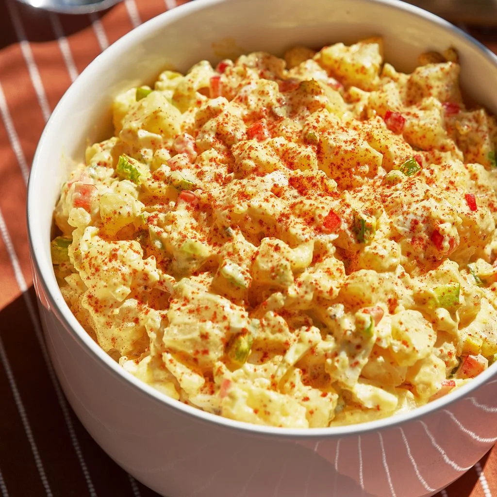 A serving bowl of creamy potato salad bright yellow with a sprinkling of paprika across the top.