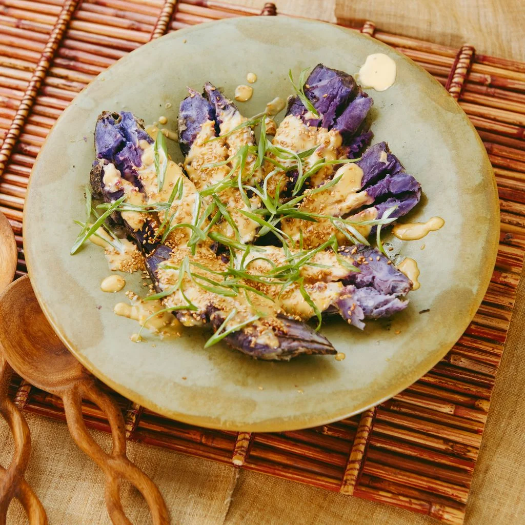 A plate of grilled purple sweet potatoes garnished with a yellow dressing thinly sliced scallions and sesame seeds.