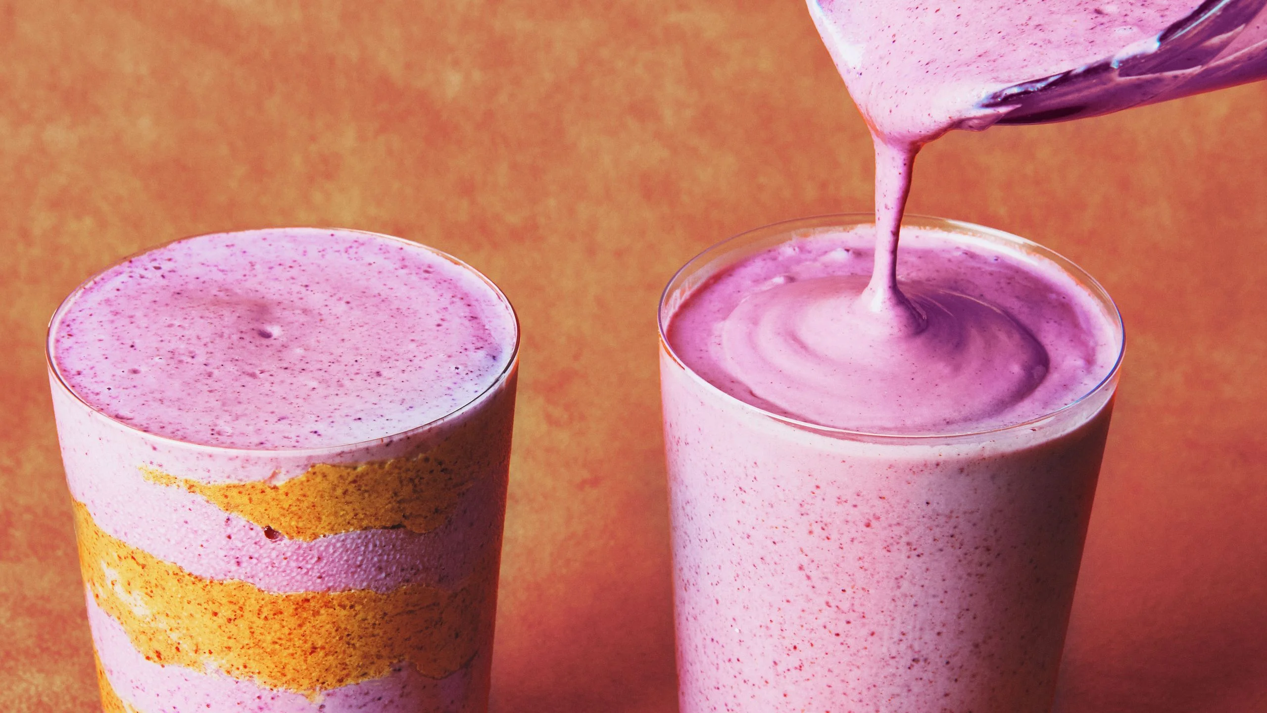 Blended mixed berries cottage cheese water and almond butter being poured into two glass cups.