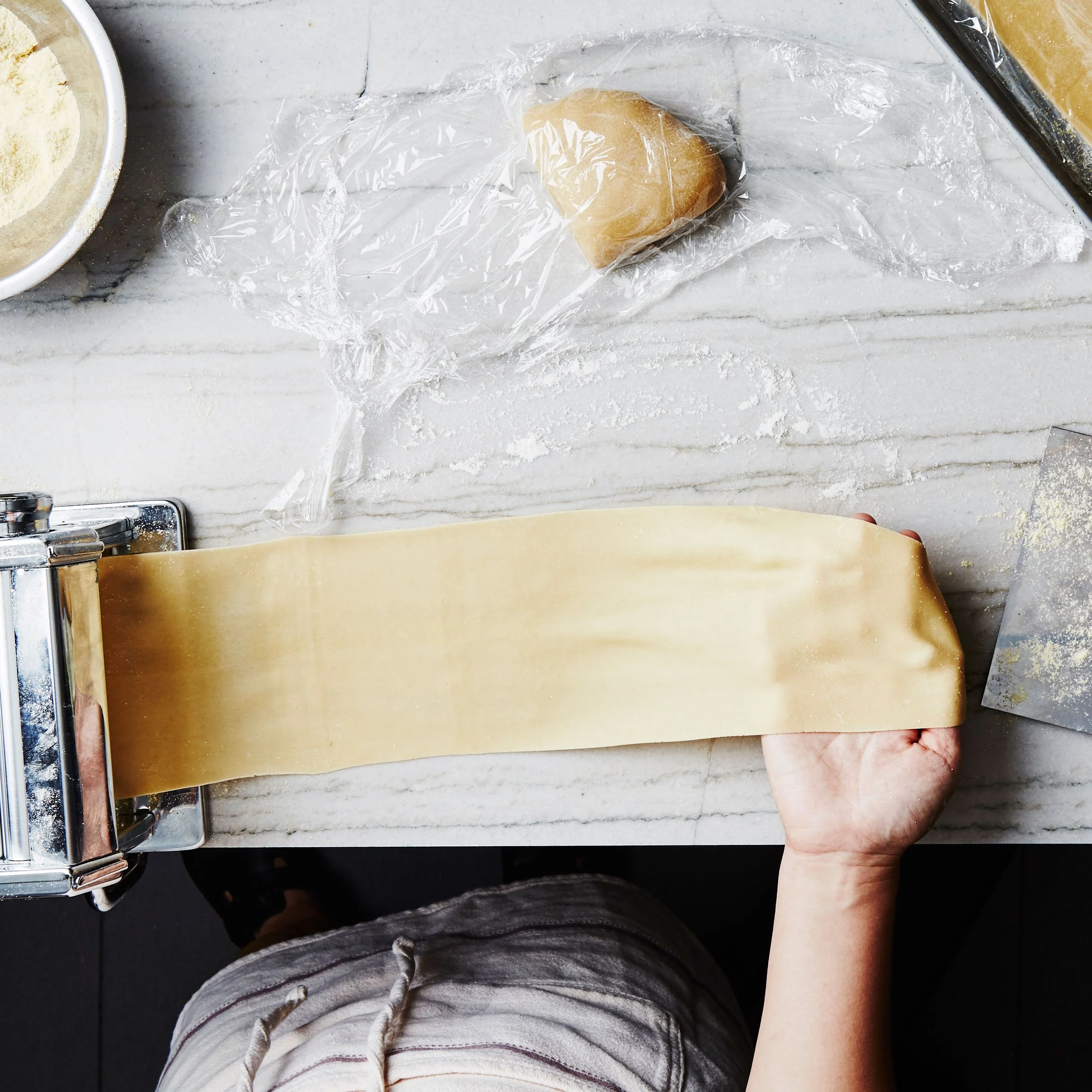 Homemade pasta dough being rolled into thin sheets with a handcrank pasta machine.
