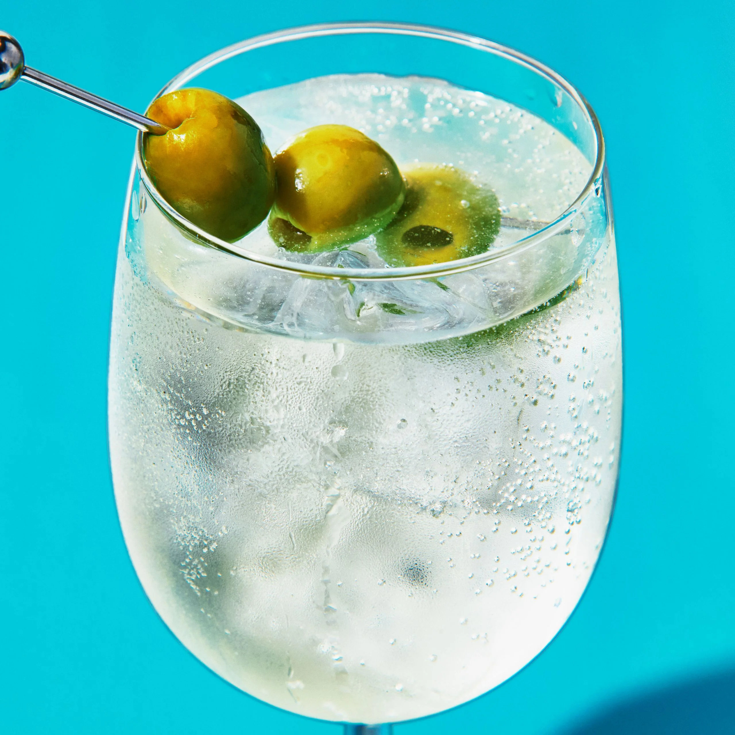 A bubbly dirty martini spritz served over ice in a stemmed wine glass and garnished with three Castelvetrano olives on a...