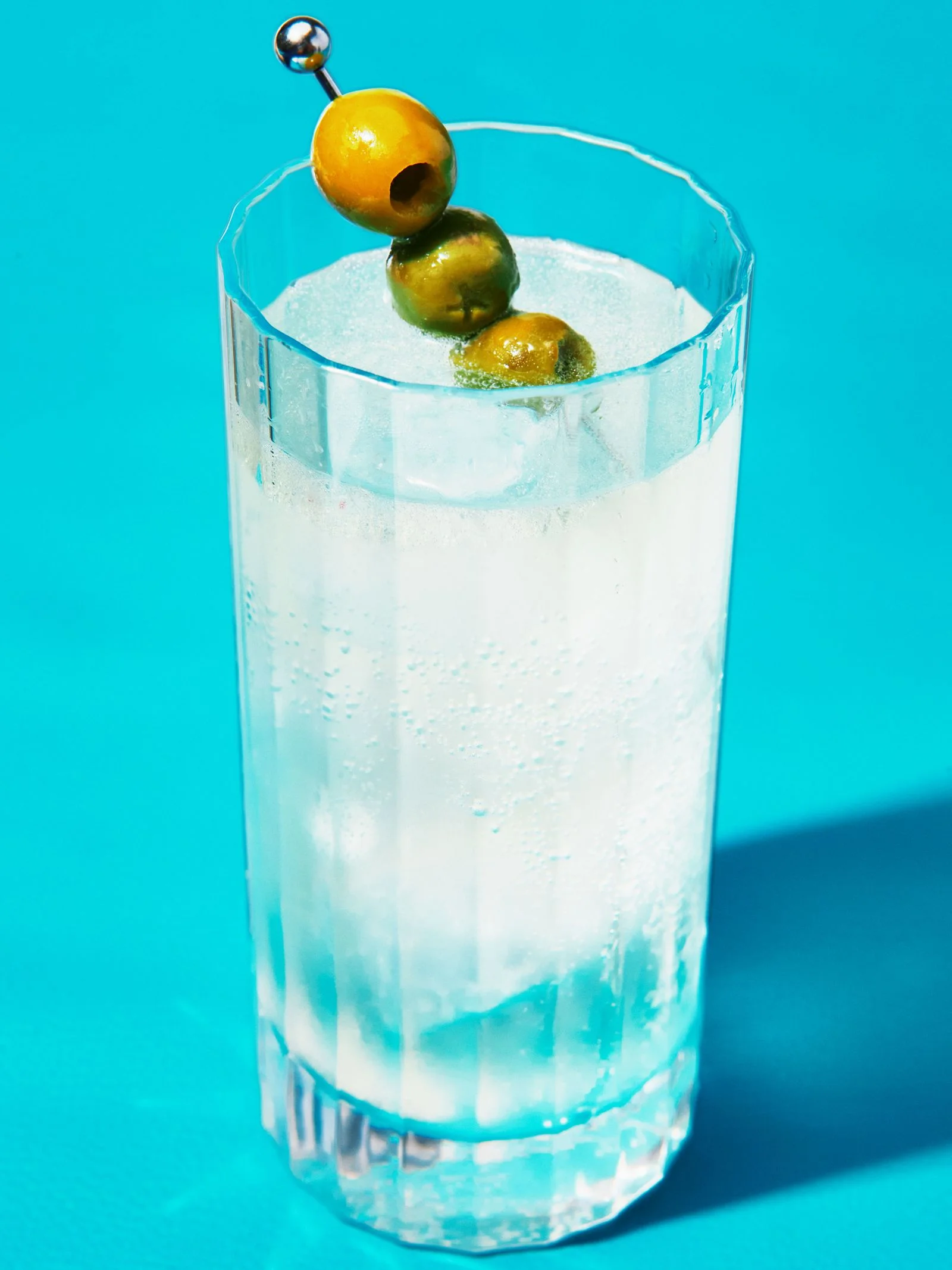 A dirty martini spritz served over ice in a Collins glass and garnished with three Castelvetrano olives on a cocktail pick.