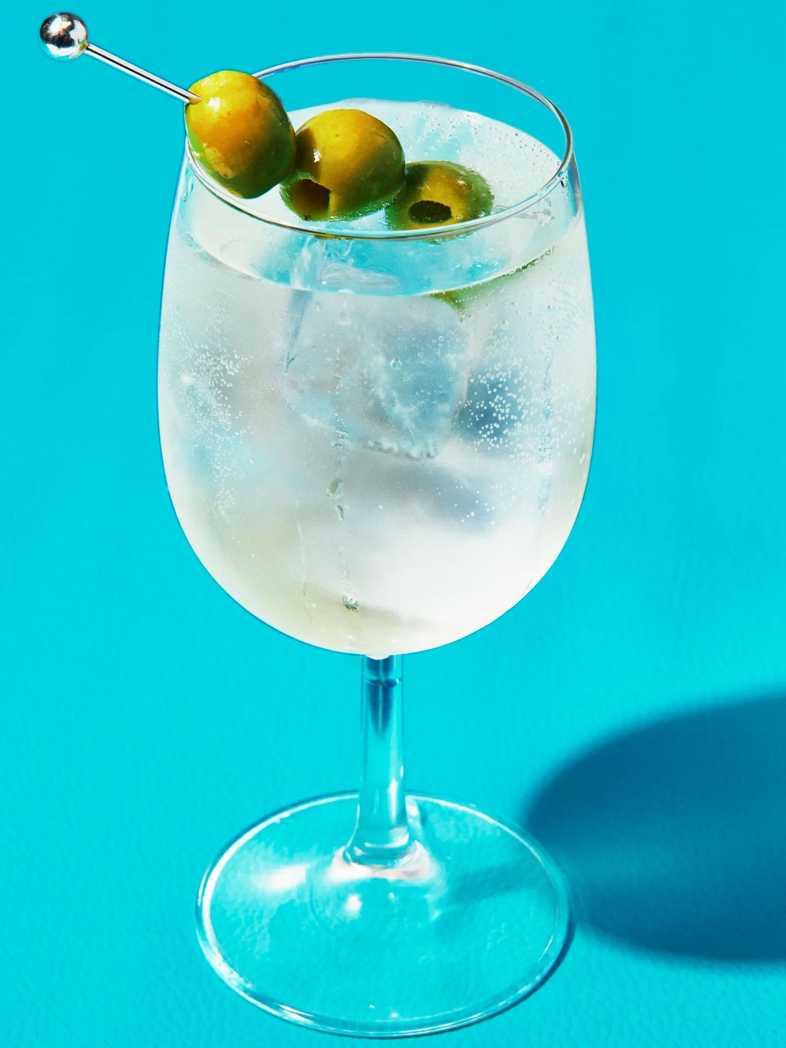 A dirty martini spritz served over ice in a stemmed wine glass against a bright blue background garnished with three...