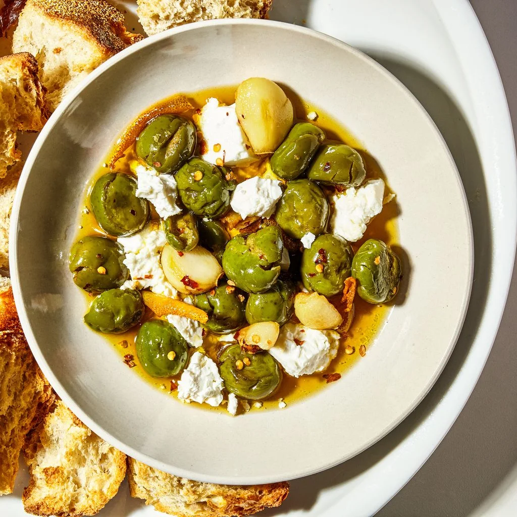 Marinated crumbles of feta cheese and Castelvetrano olives in a bowl with garlic and red pepper flakes alongside cubes...