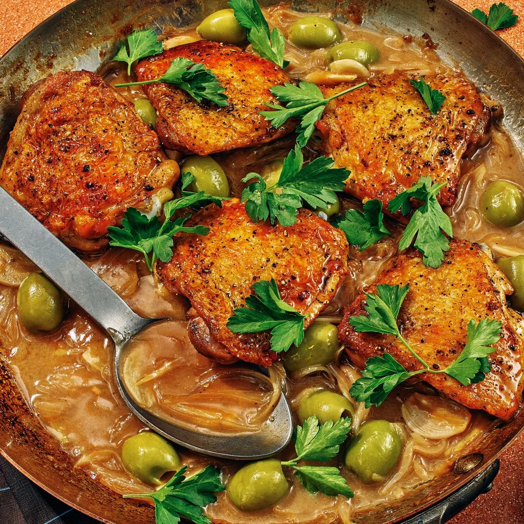 Goldenbrown chicken thighs in a skillet with green olives onions parsley and lemon wedges.