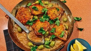 Goldenbrown chicken thighs in a skillet with green olives onions parsley and lemon wedges.