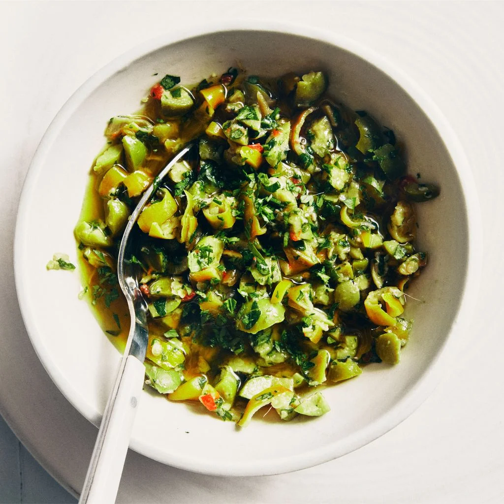 Chopped green olives pickled peppers parsley anchovies garlic red pepper flakes and olive oil in a bowl with a spoon.