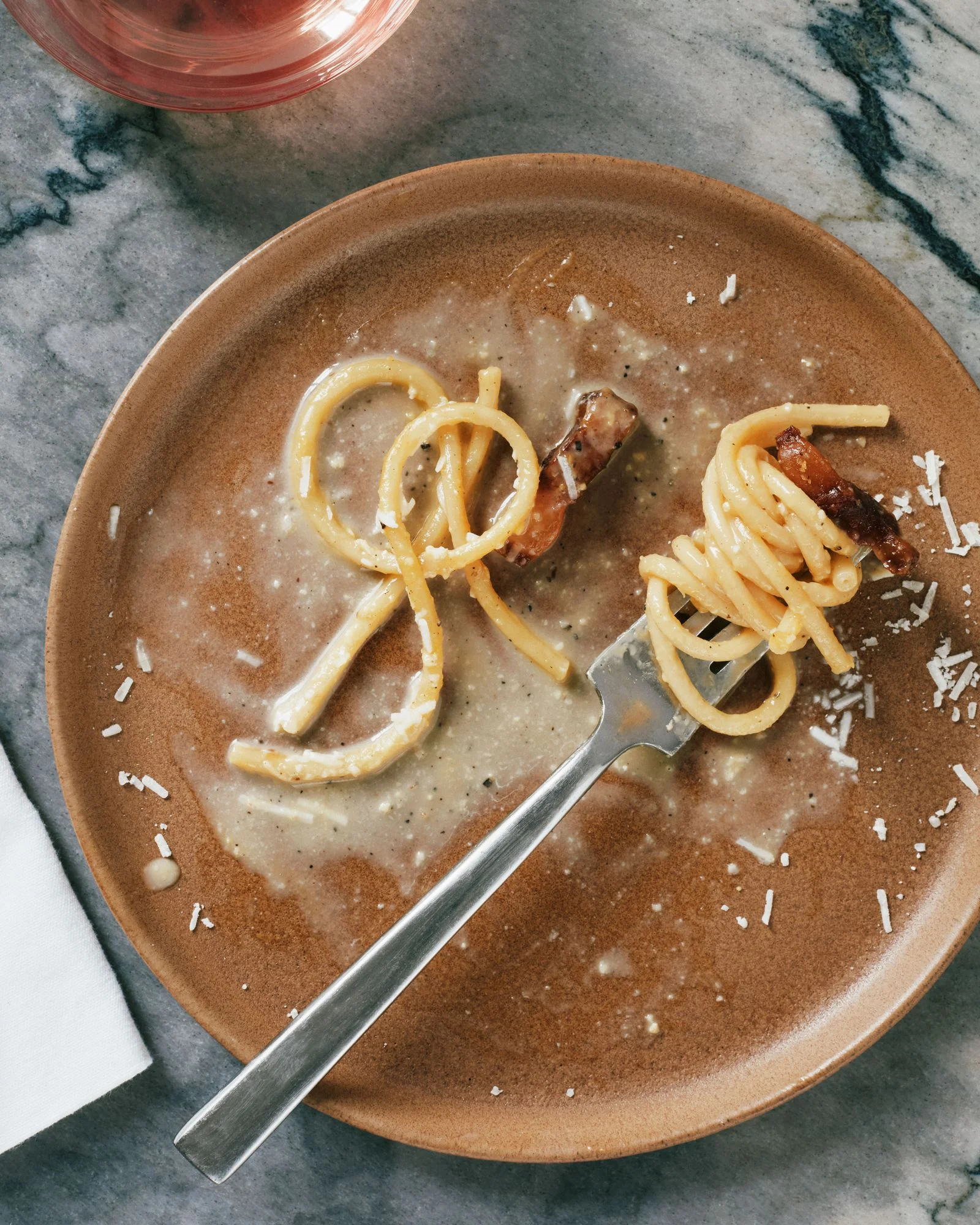 A nearempty plate of spaghetti carbonara with guanciale and shredded Pecorino with the pasta twirled around a fork.