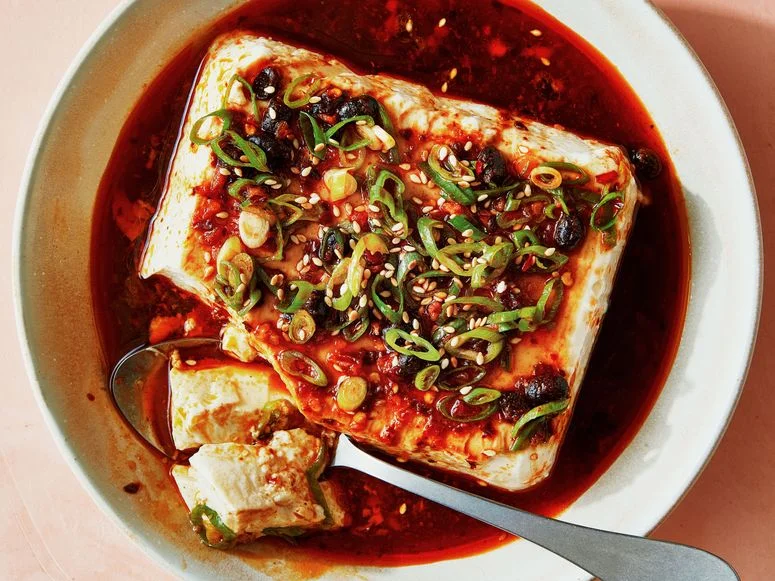 A block of silken tofu in a shallow bowl topped with chili crisp and sliced scallions.