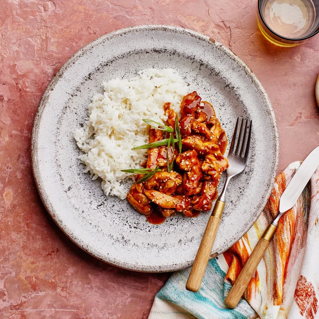 A stir fry of chicken teriyaki on a plate with steamed white rice garnished with sesame seeds and sliced scallion greens.