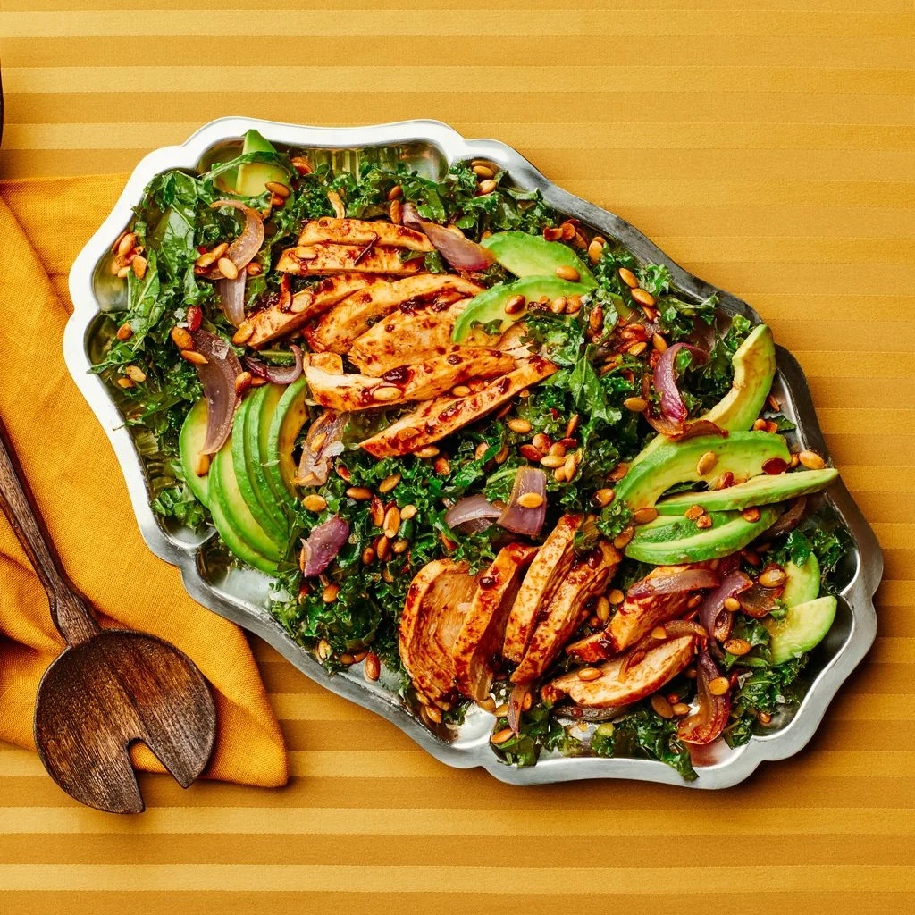 Kale salad with sliced chipotlerubbed chicken and sliced avocado on a metal platter.
