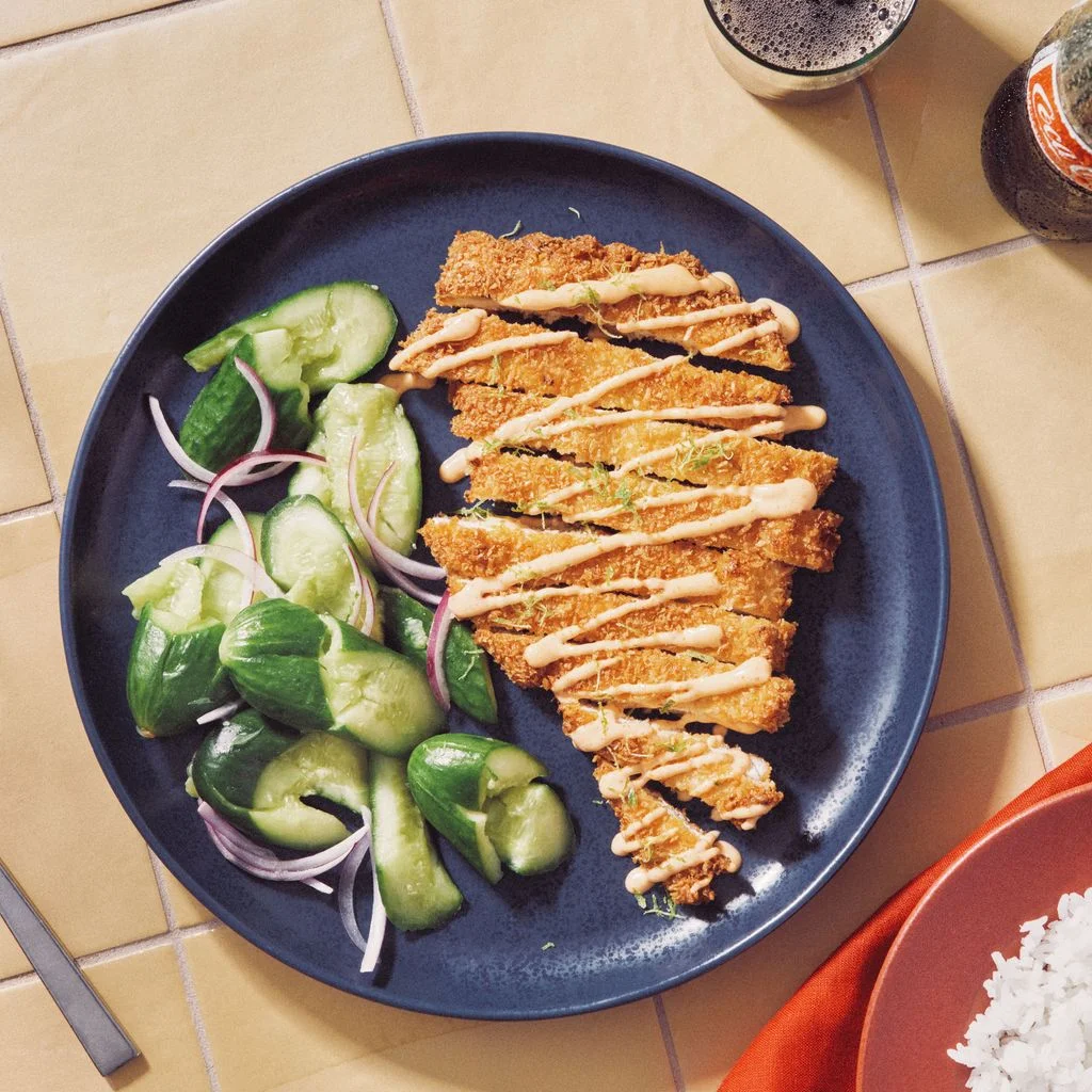 Sliced fried chicken in a pankococonut mixture topped with sriracha mayo and lime zest on a plate with smashed Persian...