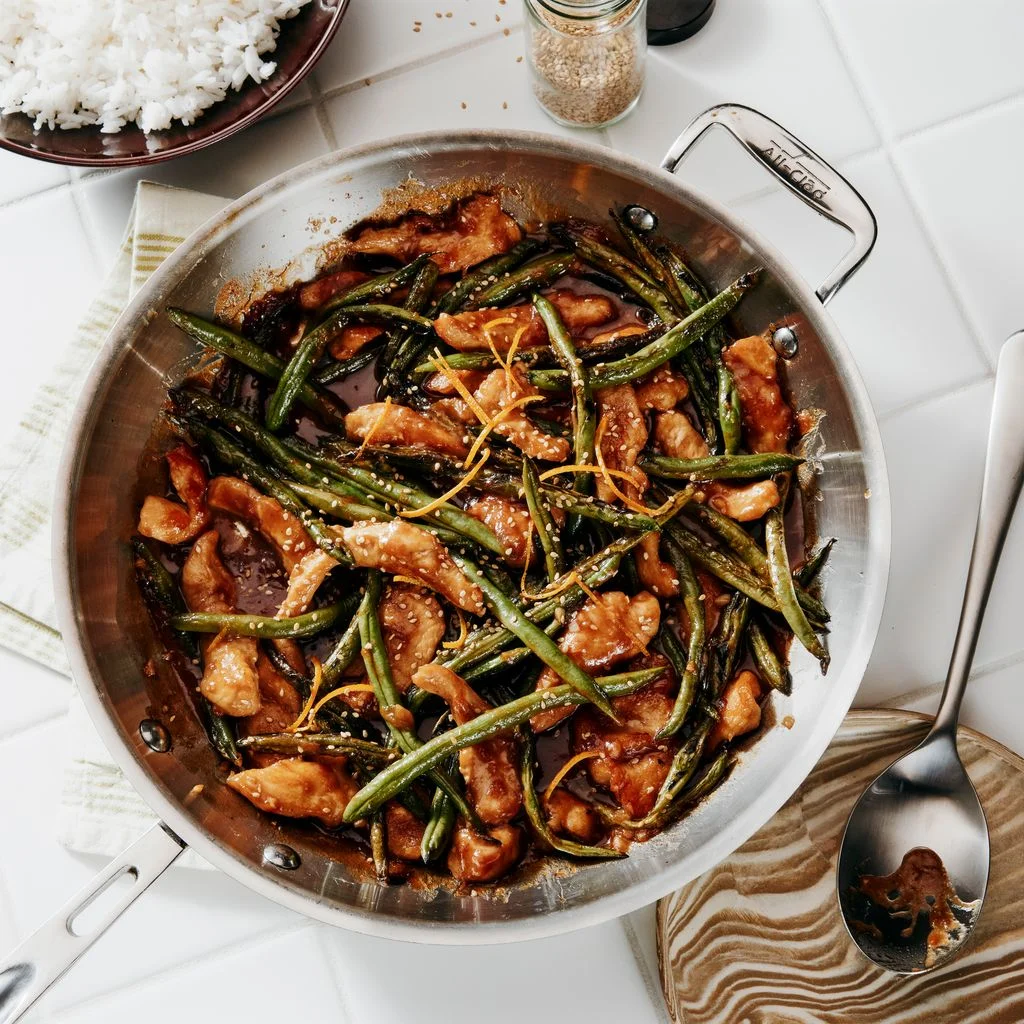 A stainless steel skillet of sweet and sour cranberry chicken and green beans atop a white tiled surface and alongside a...