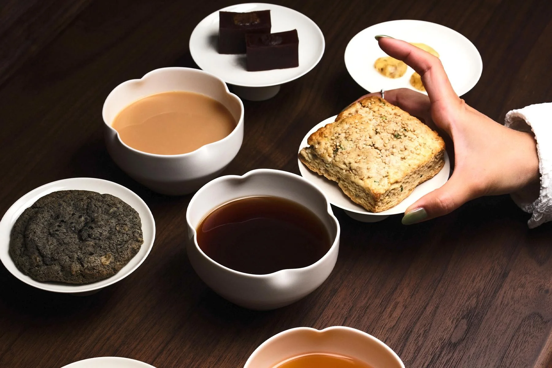 an angled shot showing a selection of tea in porcelain vessels at the manhattan teahouse soft hours. plates of confections sit next to them and a hand is holding up one plate with a baked good.