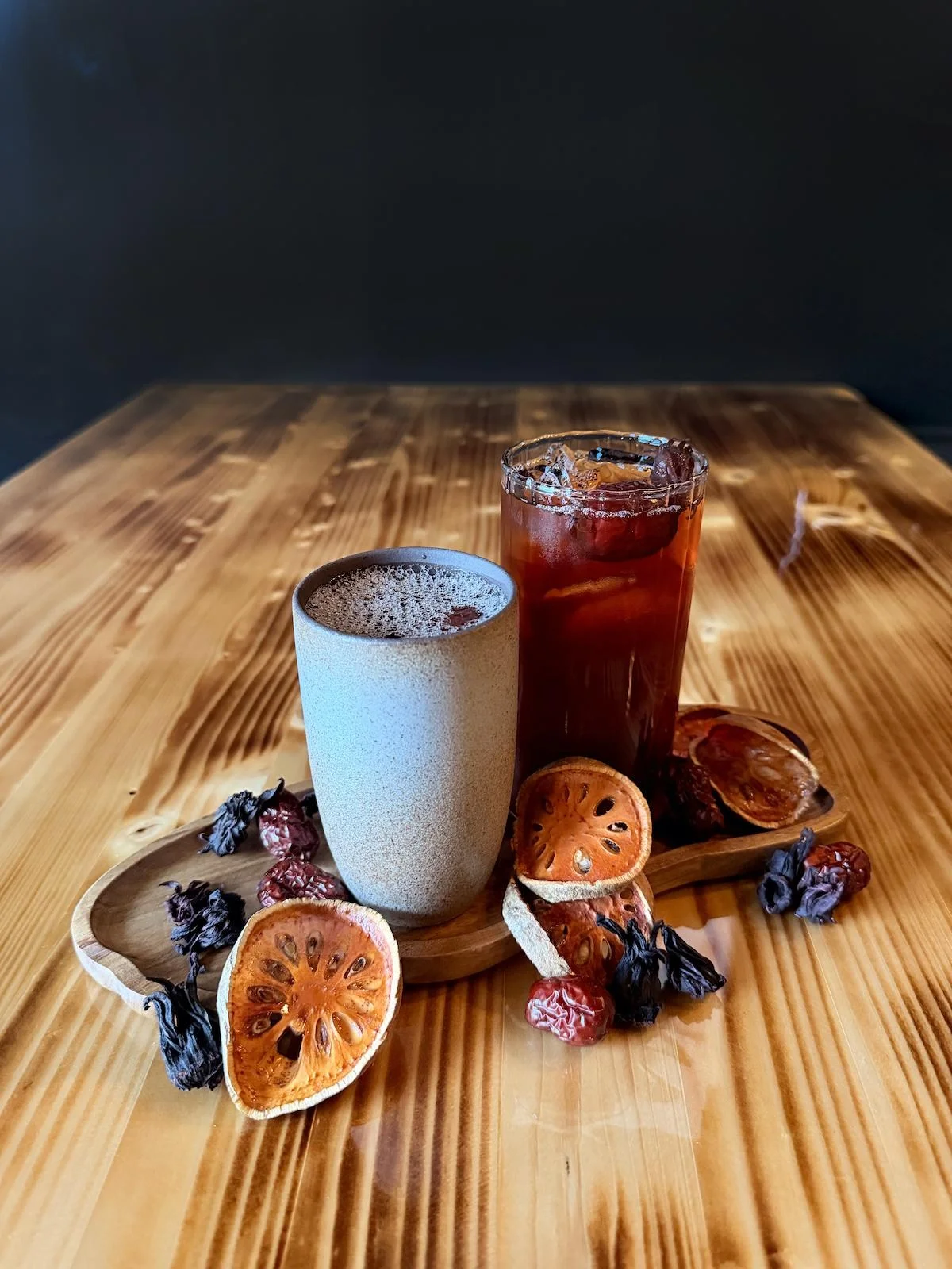 two glasses of iced tea sit on a wooden table. they’re on a tray that is also holding dried hibiscus flowers and dried slices of the fruit known as bael, also known as stone apple or golden apple. the slices are orange with seeds in a pattern going around the center point.