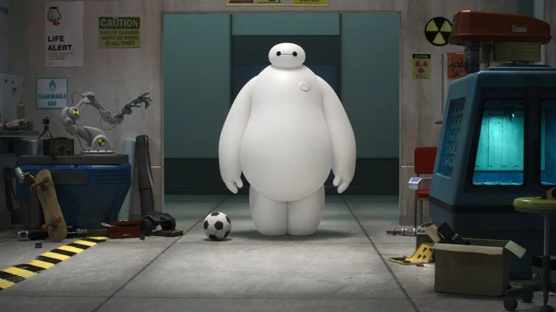 Scott Adsit as Baymax, standing straight up with his arms at his sides in a room with various objects around.