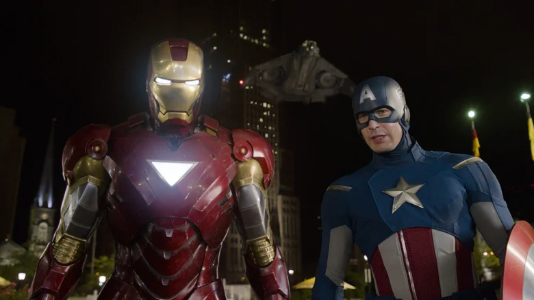 Robert Downey Jr. as Tony Stark/Iron Man and Chris Evans as Steve Rogers/Captain America in The Avengers, standing next to each other in costume at night in front of a tall building and flying ship.