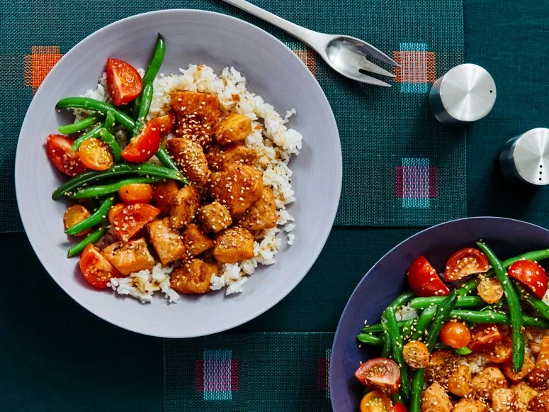 Two bowls in different hues of purple filled with cubes of soyglazed salmon atop white rice and alongside string beans...