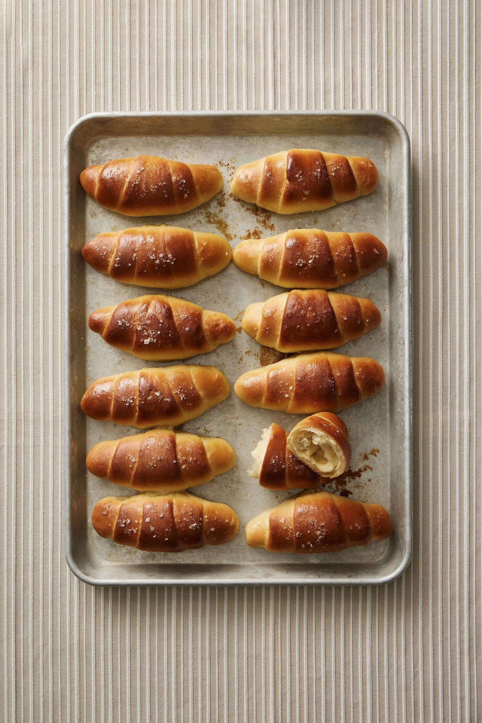 Salt Bread rolls line a metal baking sheet in two columns with coarse salt on top and one roll cut open.
