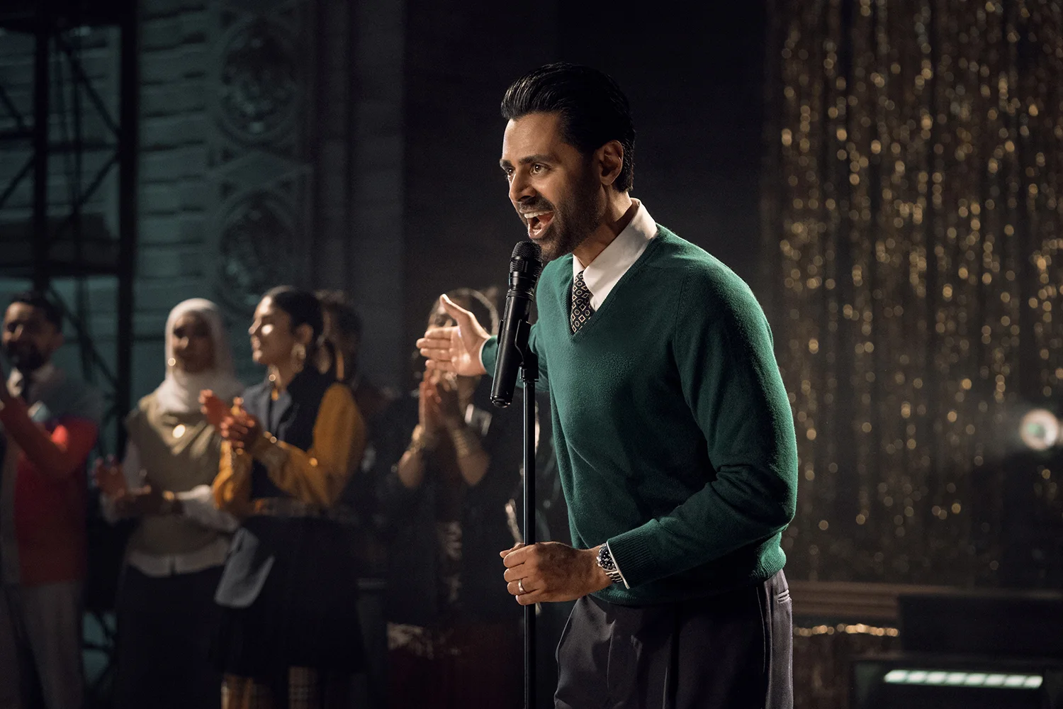 BOB 20250801 0320 R "Man in green sweater speaks into microphone on stage, with an audience of people clapping and watching in a warmly lit, festive indoor environment with golden decorations in the background."