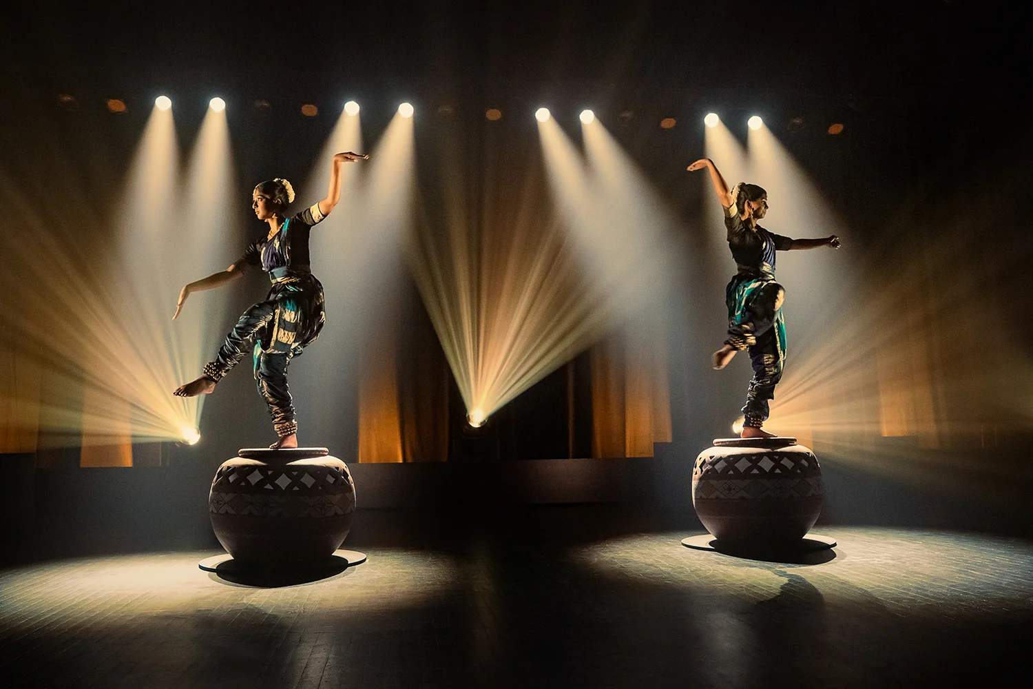 BOB 20250808 0286 R "Two performers in traditional attire balancing on large decorated drums on a stage, dramatic spotlights and beams illuminating them in a dark theater setting."